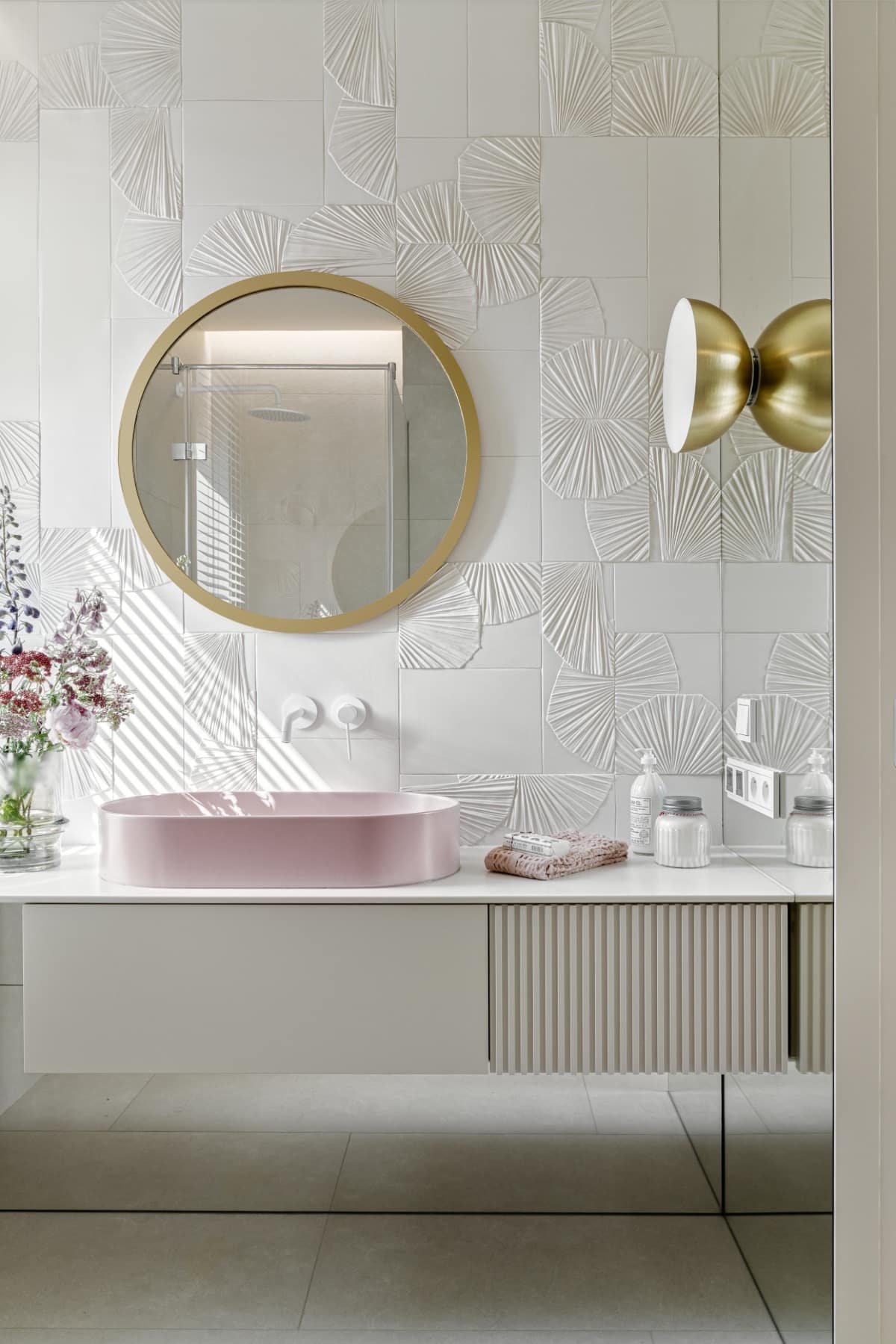 Een moderne badkamer met een witte wastafel, een ronde gouden spiegel, en een muur versierd met witte tegels in een abstract patroon, met accenten van goudkleurige armaturen en bloemen op het aanrecht.